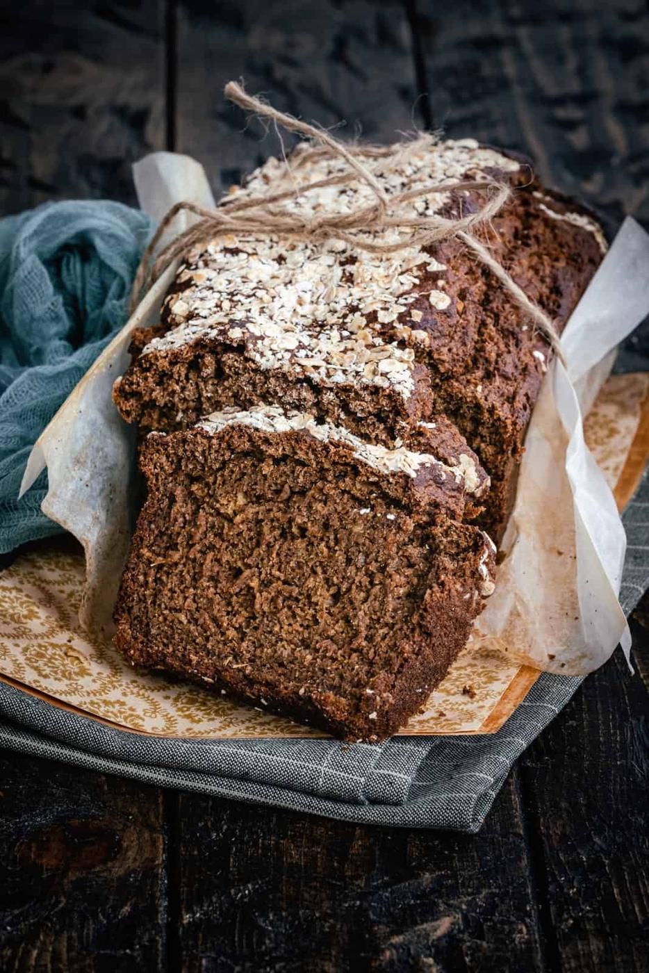 A loaf of dark brown Guiness Bread, partially sliced, sits on parchment paper atop a wooden board. The bread is topped with oats and tied with rustic twine. A blue cloth is placed beside it on a dark wooden surface.