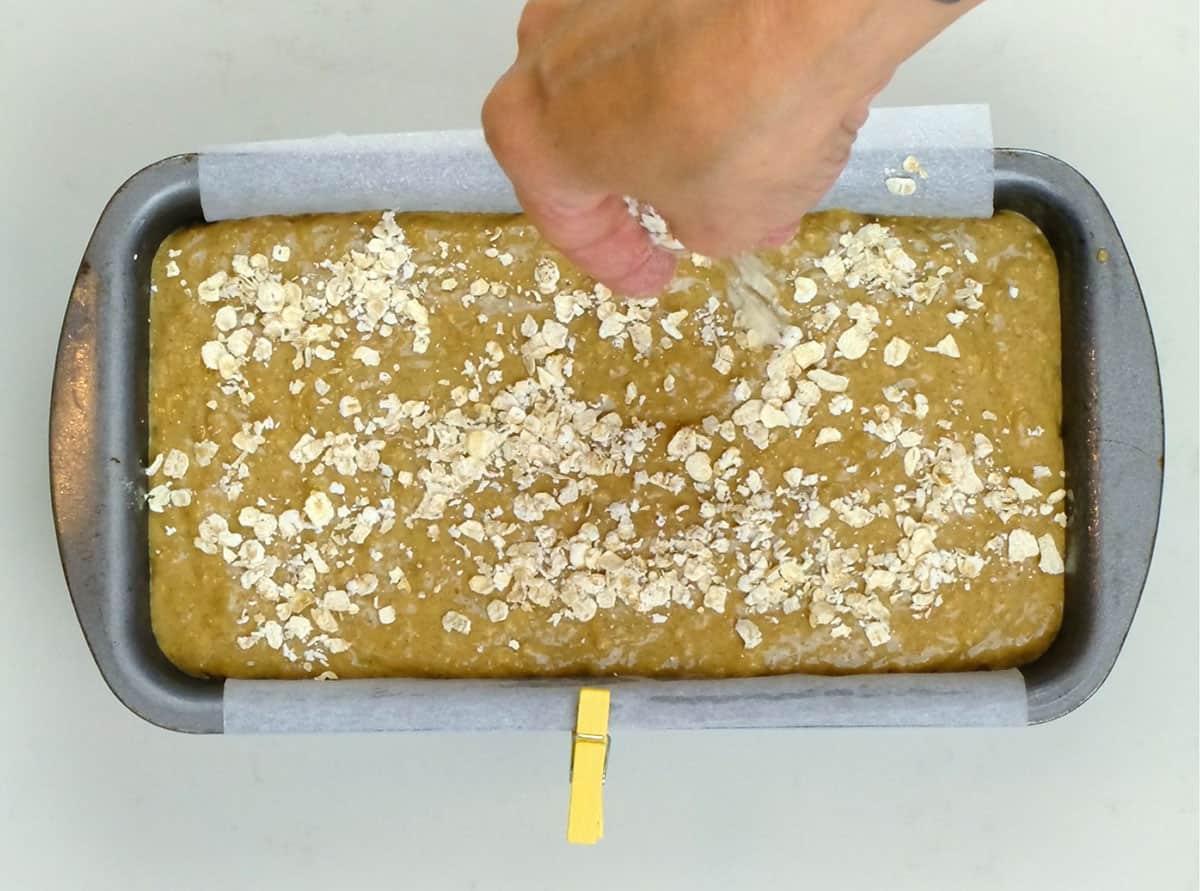 A hand sprinkles oats on top of unbaked batter in a parchment-lined loaf pan, preparing a classic Irish brown bread recipe for baking.