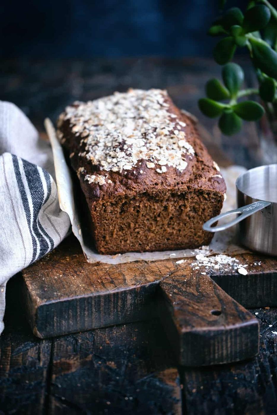 A loaf of dark, rustic Guinness Bread topped with oats sits on a wooden cutting board, next to a striped cloth and a metal measuring cup with oats. A green plant is partially visible in the background.