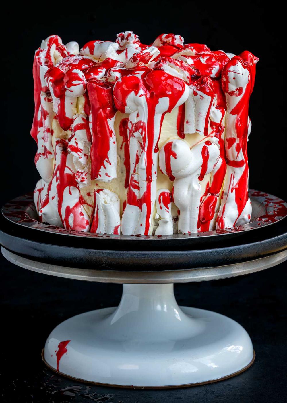 A white cake decorated with meringue bones and drizzled with bright edible fake blood on a pedestal stand.