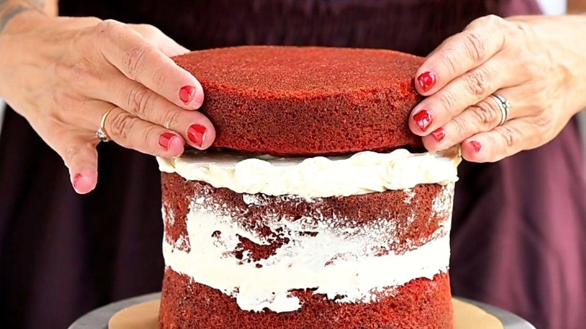 Hands place a red velvet cake layer on top of a frosted stack decorated with meringue bones.