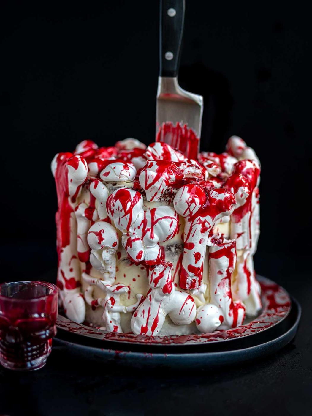 A cake covered in white meringue bones and edible fake blood drips, with a large knife stuck into the top and a small skull-shaped glass of red liquid beside it.