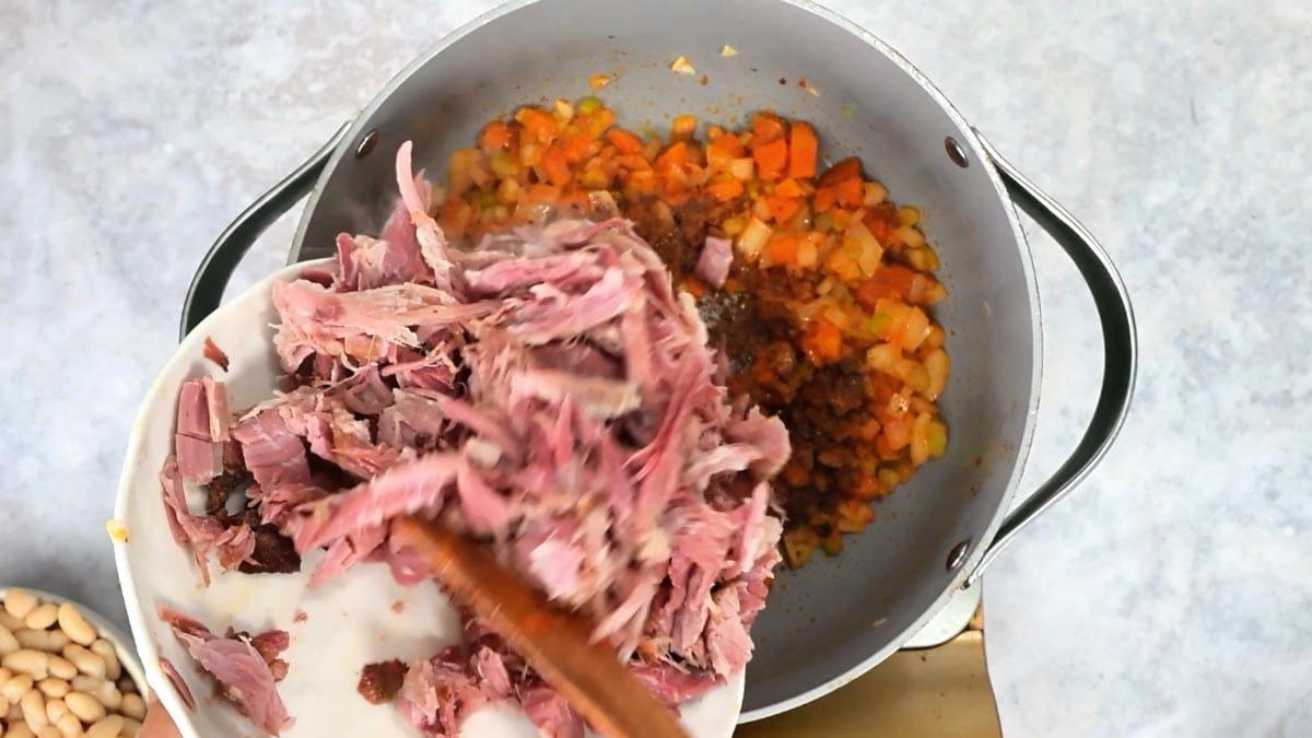 Shredded cooked ham being added from a plate to the pot of softened carrots, onion and celery for ham and bean soup.