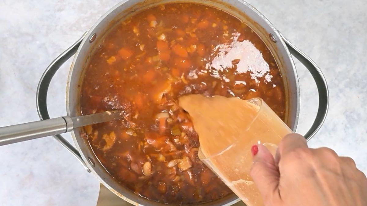 A hand pouring broth from a measuring cup into a large pot of ham and bean soup.