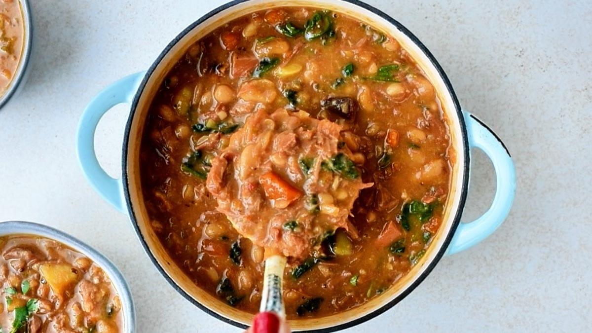 A ladle lifts a scoop of hearty ham and bean soup from a blue enameled pot.