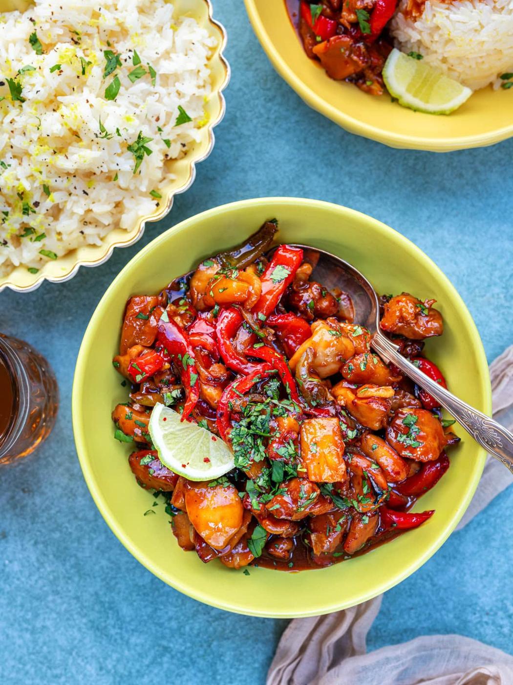 A bowl of Hawaiian pineapple chicken with colorful bell peppers and sauce, garnished with chopped herbs and a lime slice, sits next to a bowl of rice and a small dish of sauce on a blue surface.
