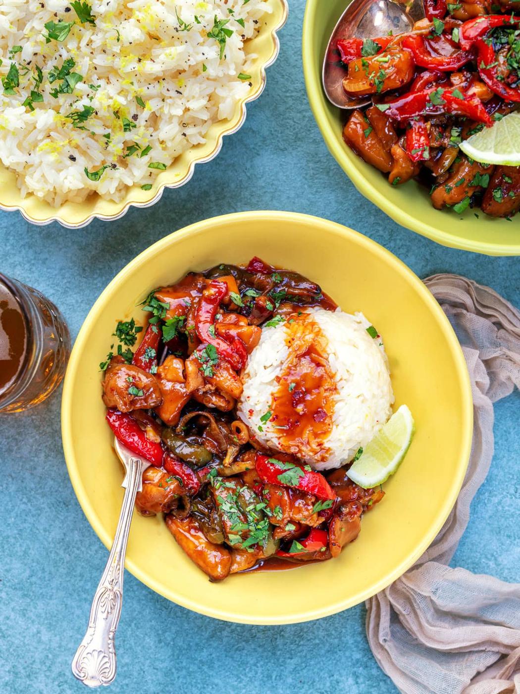 A yellow bowl with pineapple chicken, red bell peppers, and vegetables in sauce, served with a mound of white rice and a lime wedge. Nearby are a bowl of rice, another dish of stir-fry, and a drink on a blue surface.