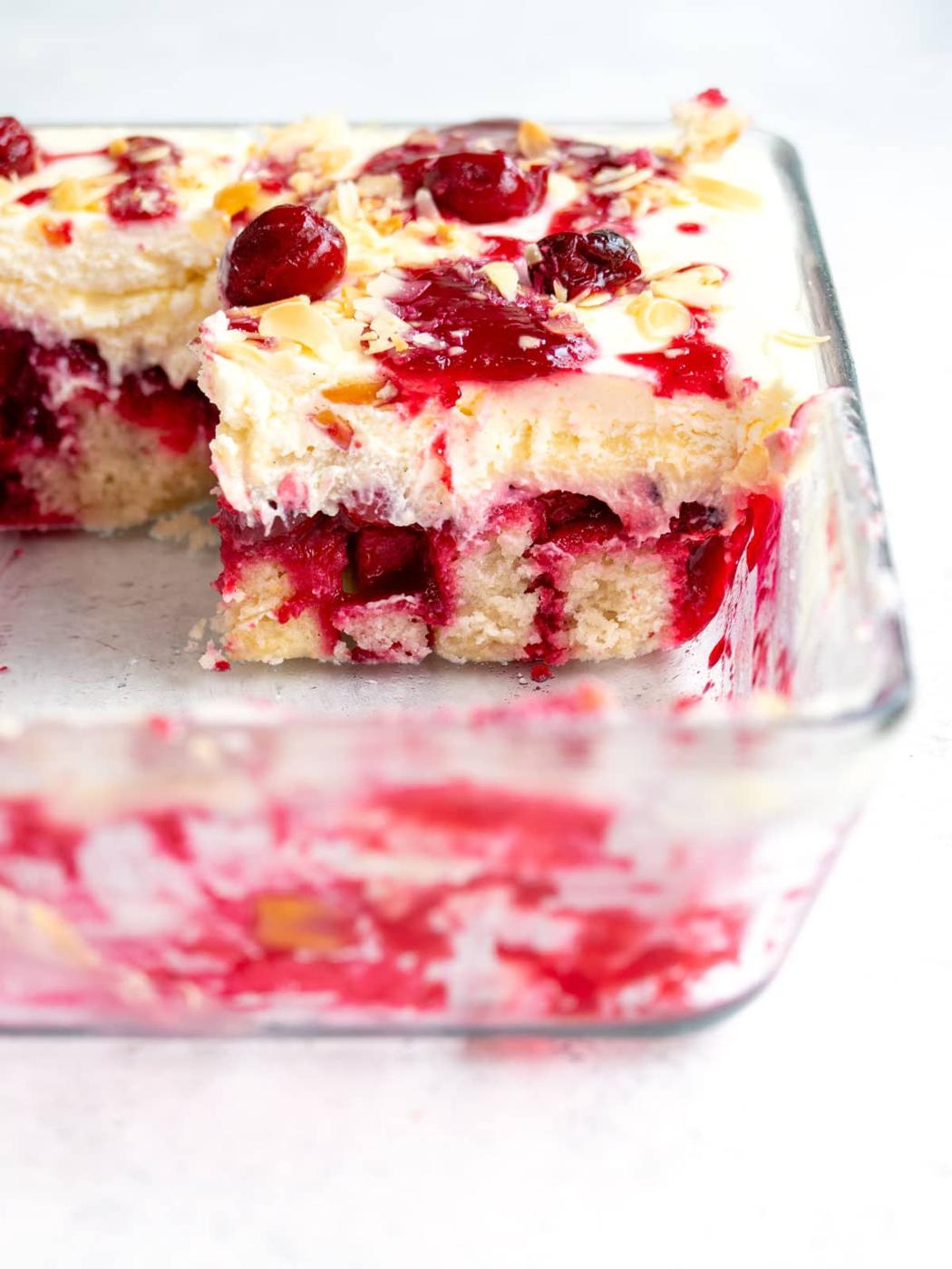 A glass baking dish containing partially eaten cherry almond cake with visible layers of cake, cream, and cherry topping, garnished with sliced almonds and whole cherries.