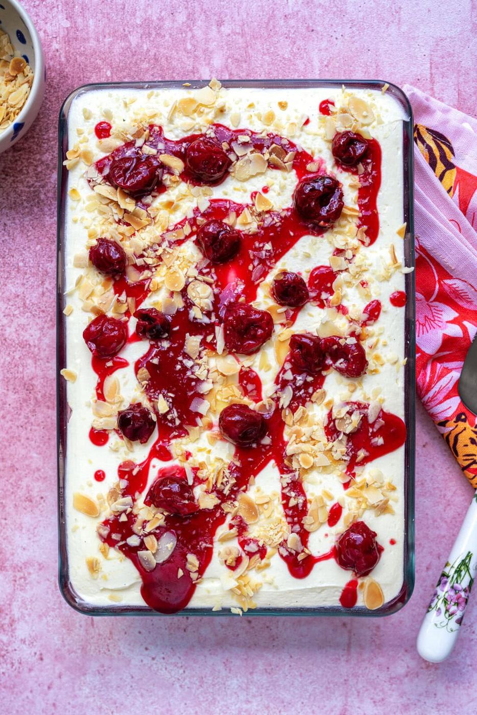 A rectangular glass dish filled with a creamy dessert, topped with cherry sauce, whole cherries, and sliced almonds, on a pink surface with a colorful napkin and a white-handled utensil beside it.