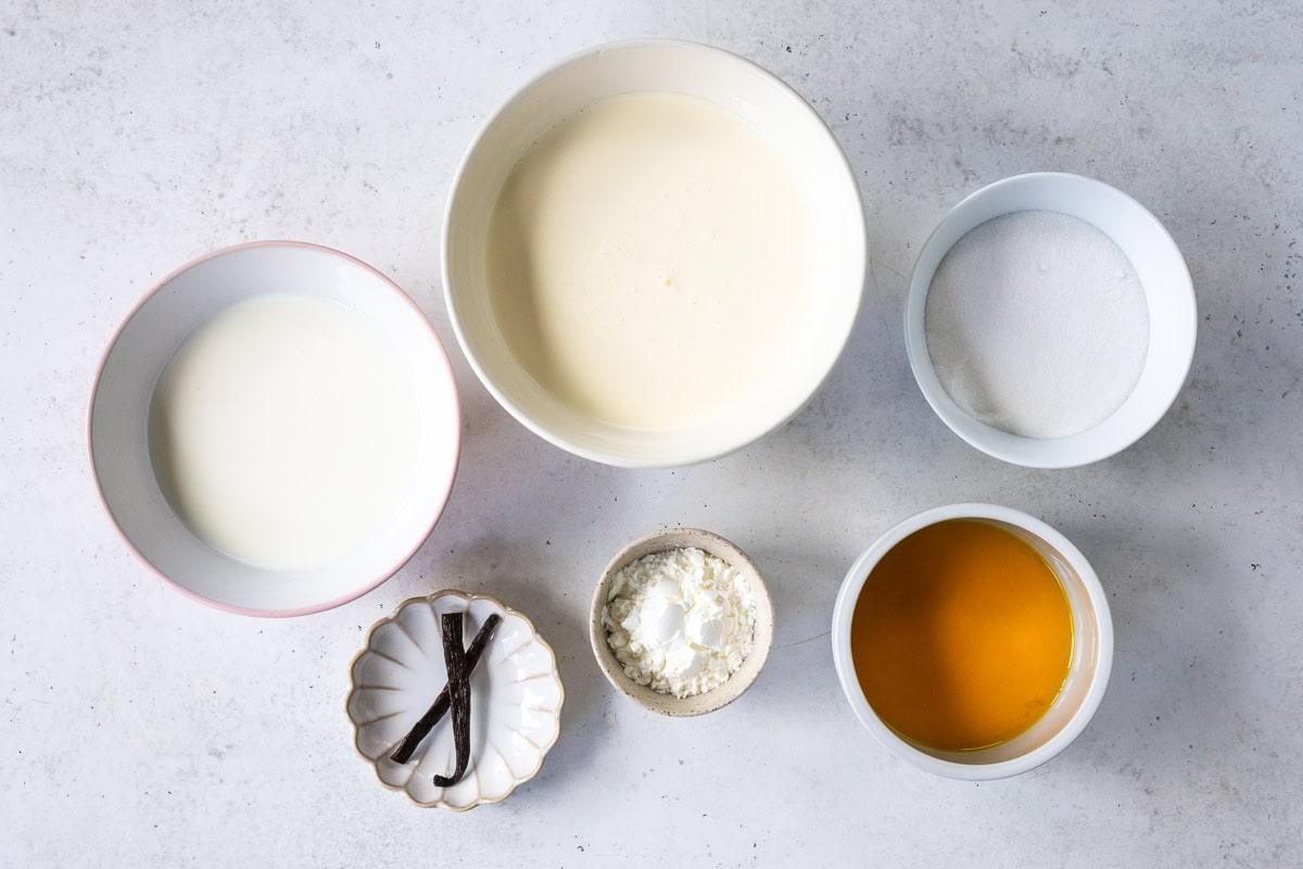 Six bowls on a light surface containing cream, sugar, milk, honey, cornstarch, and a dish with vanilla beans, arranged neatly for a recipe.