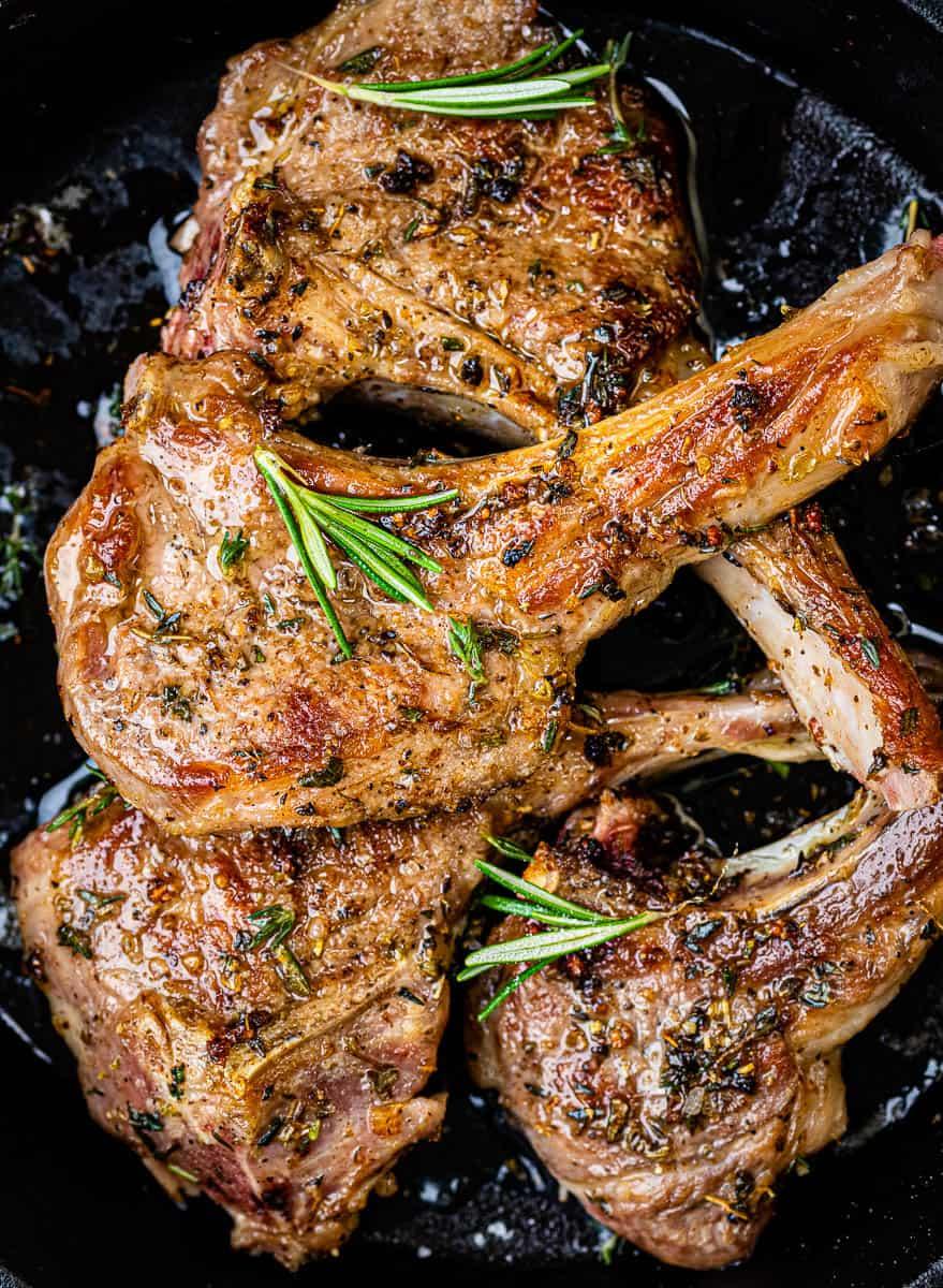 Three cooked lamb chops seasoned with herbs and garnished with fresh rosemary sprigs, resting in a dark skillet. The lamb chops appear juicy and well-seared, with visible spices and a glossy finish.