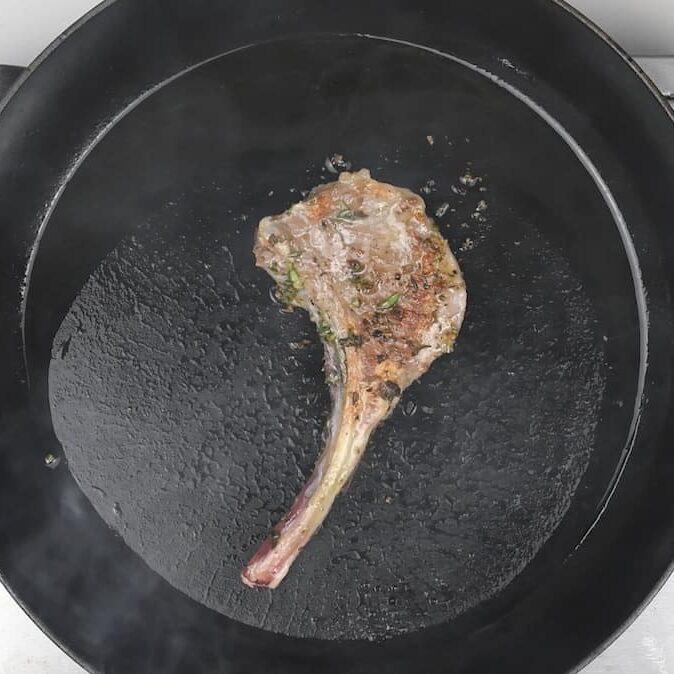 A seasoned Lamb Chop with a bone is being seared in a black skillet, surrounded by sizzling oil and fresh herbs, showcasing the rich flavors of classic Lamb Chops.