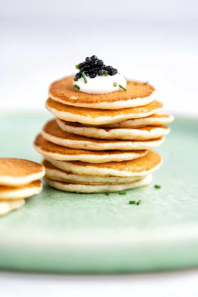 A stack of small, golden Russian pancakes is topped with a dollop of cream, black caviar, and chopped chives, placed on a light green plate—perfect for an Easy Blini Recipe.