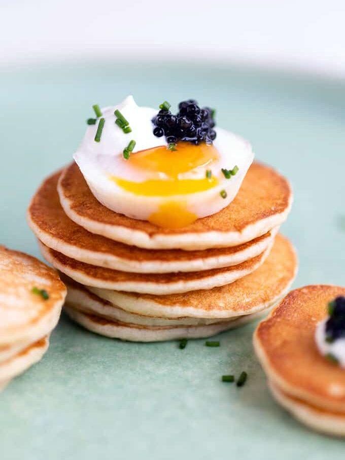 A stack of small, golden Russian pancakes topped with a soft-boiled egg, black caviar, and chopped chives is served as an elegant appetizer on a light green plate.