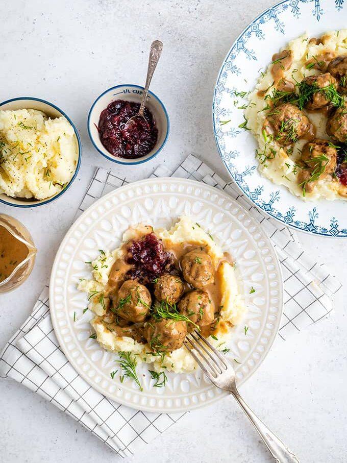 Two plates of Swedish Meatballs with creamy gravy, mashed potatoes, and lingonberry sauce are arranged on a white table with a fork. Small bowls of mashed potatoes, lingonberry sauce, and gravy are also visible.