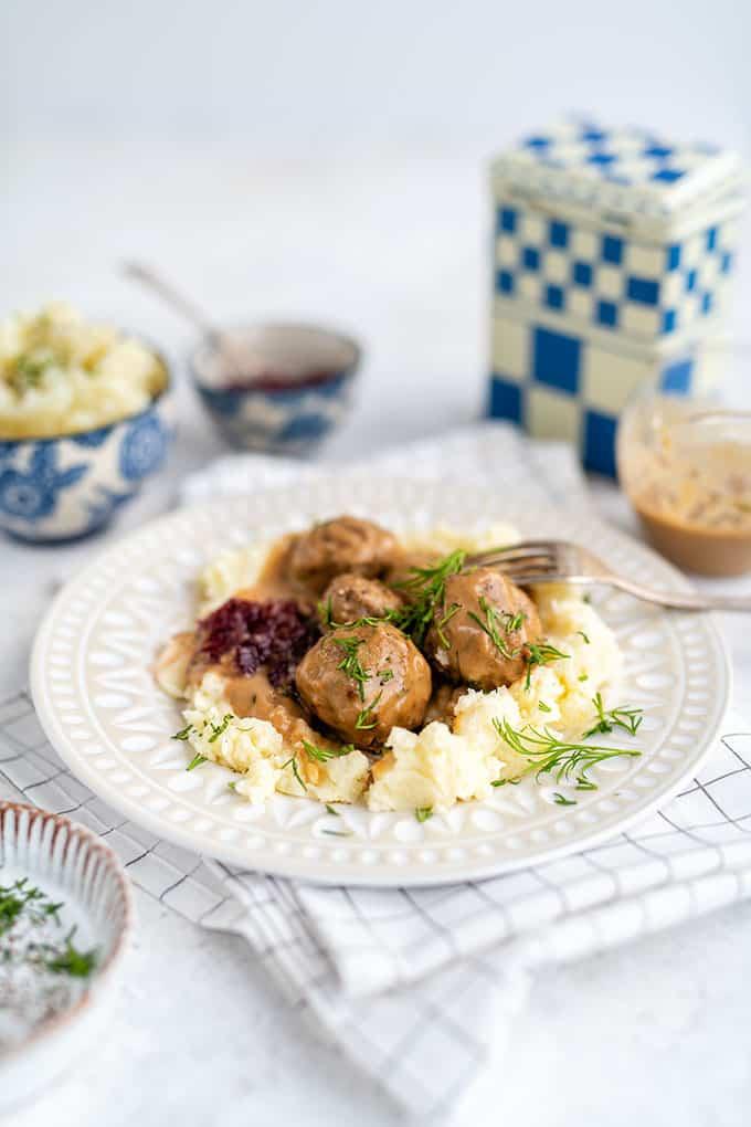A plate of Swedish meatballs served over mashed potatoes with creamy gravy, garnished with fresh dill, and a side of lingonberry sauce. The background includes small bowls and a checkered container.