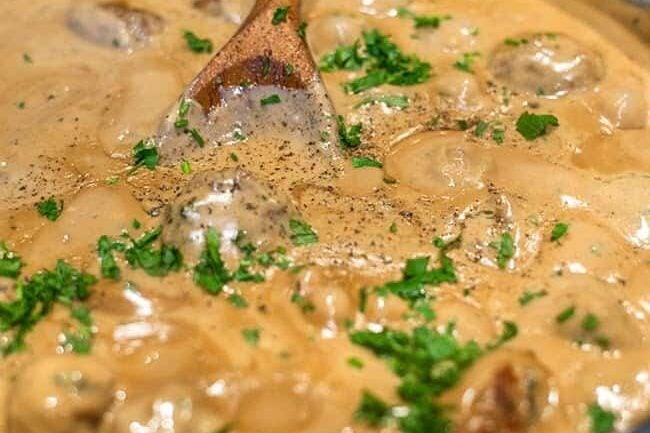 Close-up of Swedish meatballs in creamy sauce with chopped parsley on top, being stirred with a wooden spoon.