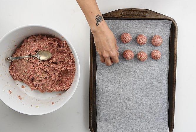 A person shapes Swedish Meatballs from a bowl of ground meat mixture and arranges them on a baking sheet lined with parchment paper. A spoon rests in the bowl, and a bee tattoo is visible on the persons wrist.