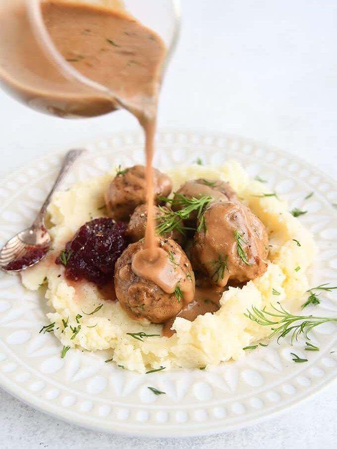 Meatballs with creamy brown gravy are served on mashed potatoes, garnished with fresh dill and a side of cranberry sauce, on a white patterned plate. Gravy is being poured over the meatballs.