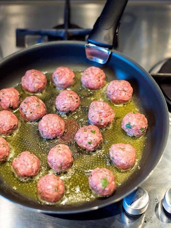 Raw meatballs are frying in a black skillet with oil on a stovetop. The meatballs are spaced evenly, starting to brown slightly, and the stove dials are visible in the foreground.