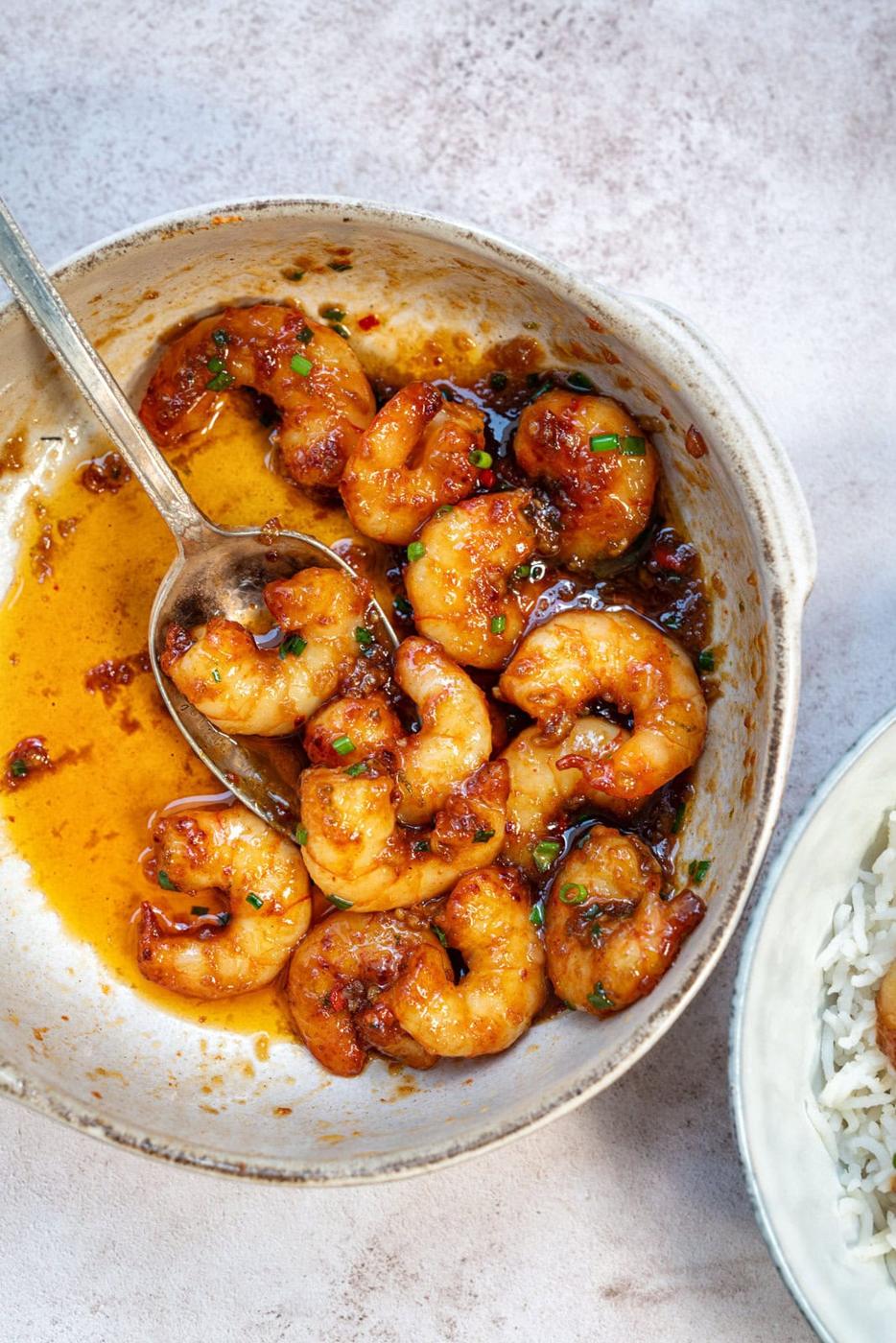 A bowl of honey garlic shrimp (prawms) in a golden, spicy sauce garnished with herbs, with a spoon resting inside. Part of a dish with white rice is visible on the side.