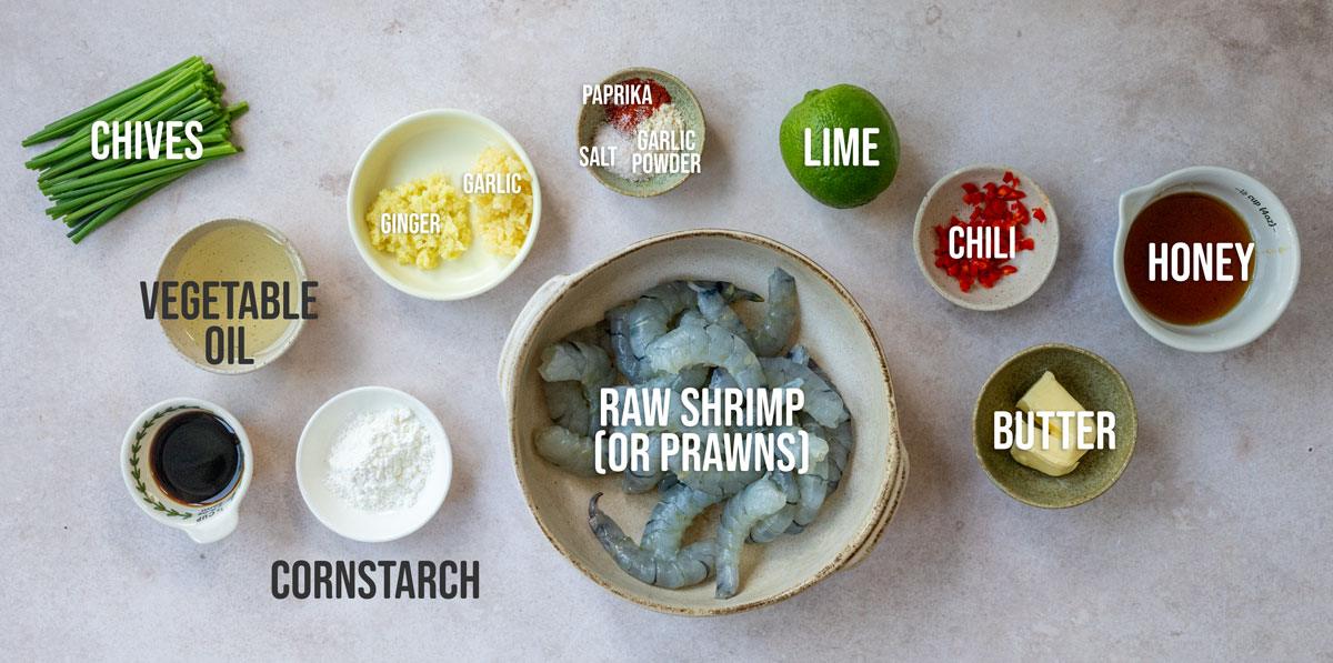 A flat lay of ingredients for a shrimp dish: chives, minced garlic, minced ginger, vegetable oil, cornstarch, soy sauce, raw shrimp, lime, paprika, salt, garlic powder, chopped chili, honey, and butter in bowls.