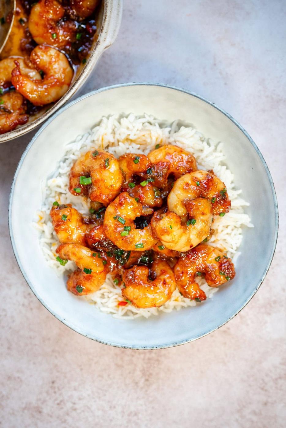 A bowl of white rice topped with saucy, glazed honey garlic shrimp garnished with chopped herbs. Another dish with more shrimp is partially visible in the corner.