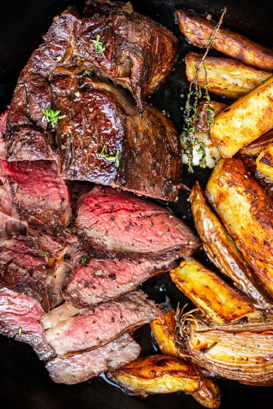Sliced medium-rare bone-in ribeye steak with a browned crust sits beside golden roasted potato wedges and sprigs of fresh thyme.