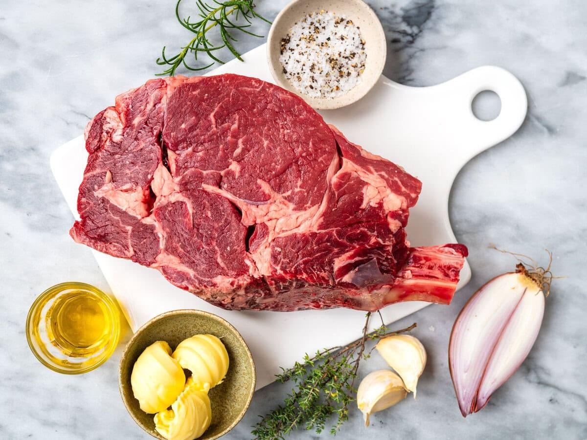 Raw ribeye steak on the bone on a white cutting board, surrounded by butter, olive oil, garlic cloves, a halved shallot, salt and pepper in a small bowl, fresh thyme, and rosemary on a marble surface.