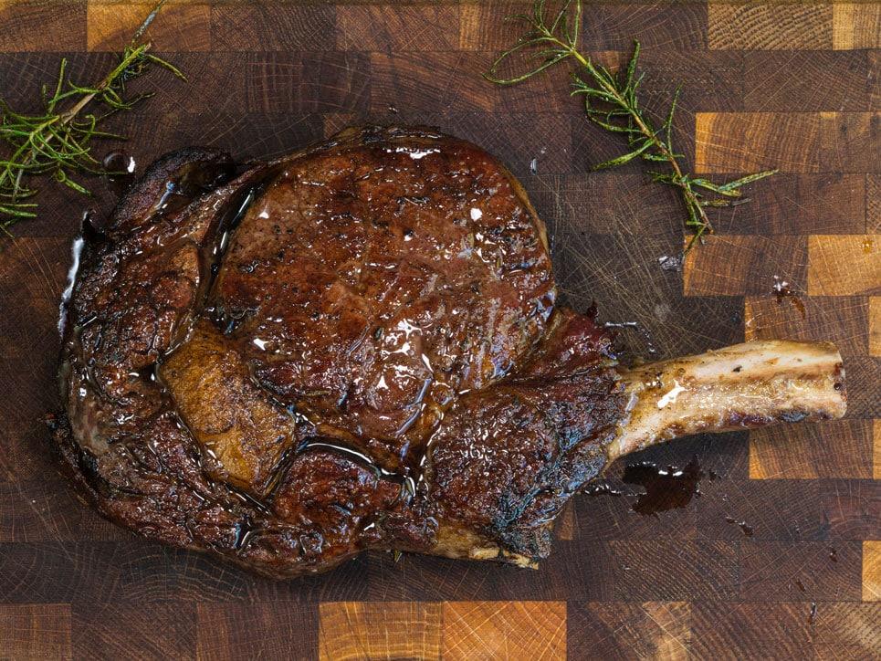 A juicy, cooked bone-in ribeye steak rests on a wooden cutting board, glistening with juices. Sprigs of fresh rosemary are placed nearby for garnish.
