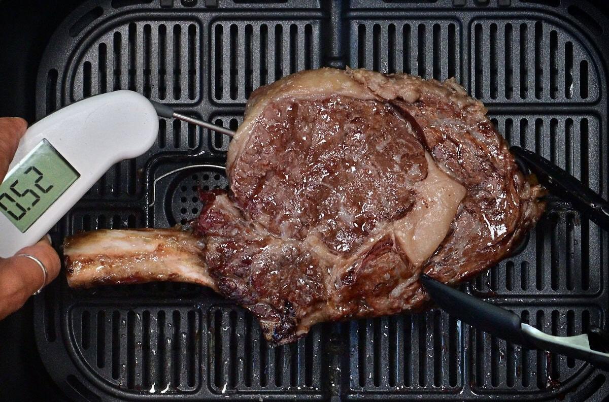 A cooked bone-in steak sits in an air fryer basket. A meat thermometer inserted into the steak reads 52°C, while a pair of tongs holds the steak in place.