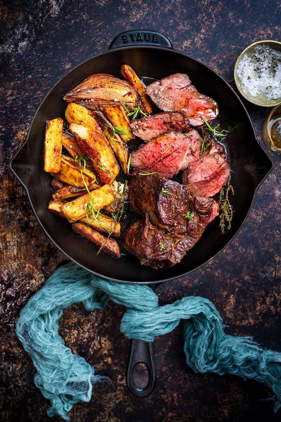 A cast iron skillet filled with sliced steak, roasted potato wedges, and caramelized onions, garnished with herbs, sits on a rustic surface next to a green cloth and a small bowl of sauce.