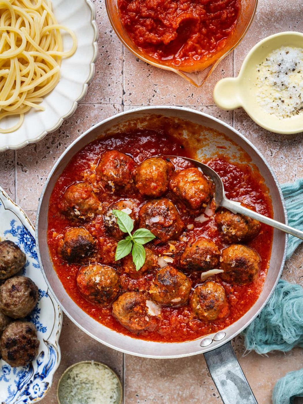 A pan of meatballs in tomato sauce garnished with basil, surrounded by a plate of spaghetti, a bowl of tomato sauce, a dish of grated cheese, and a plate with extra meatballs on a tiled surface.