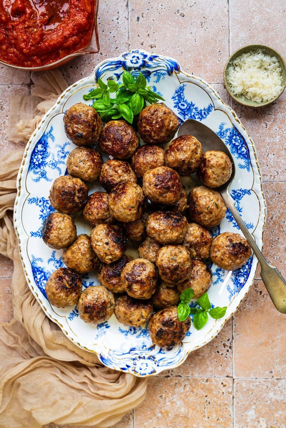 A blue and white floral plate filled with golden-brown meatballs, garnished with fresh basil, sits on a tiled surface. A spoon rests on the plate, with bowls of tomato sauce and grated cheese nearby.