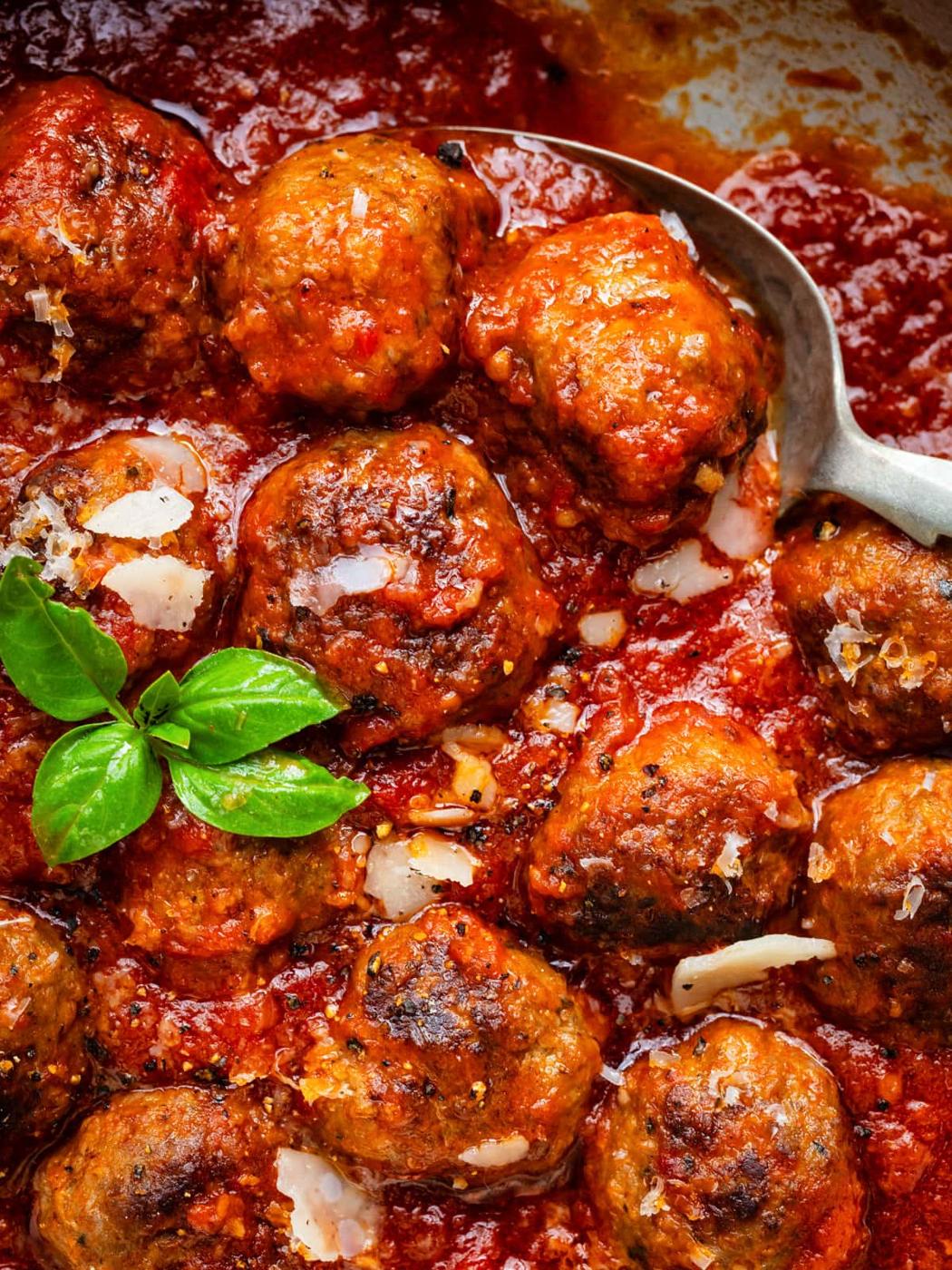 Close-up of Italian meatballs in rich tomato sauce, garnished with fresh basil leaves and parmesan shavings, with a spoon resting in the sauce.