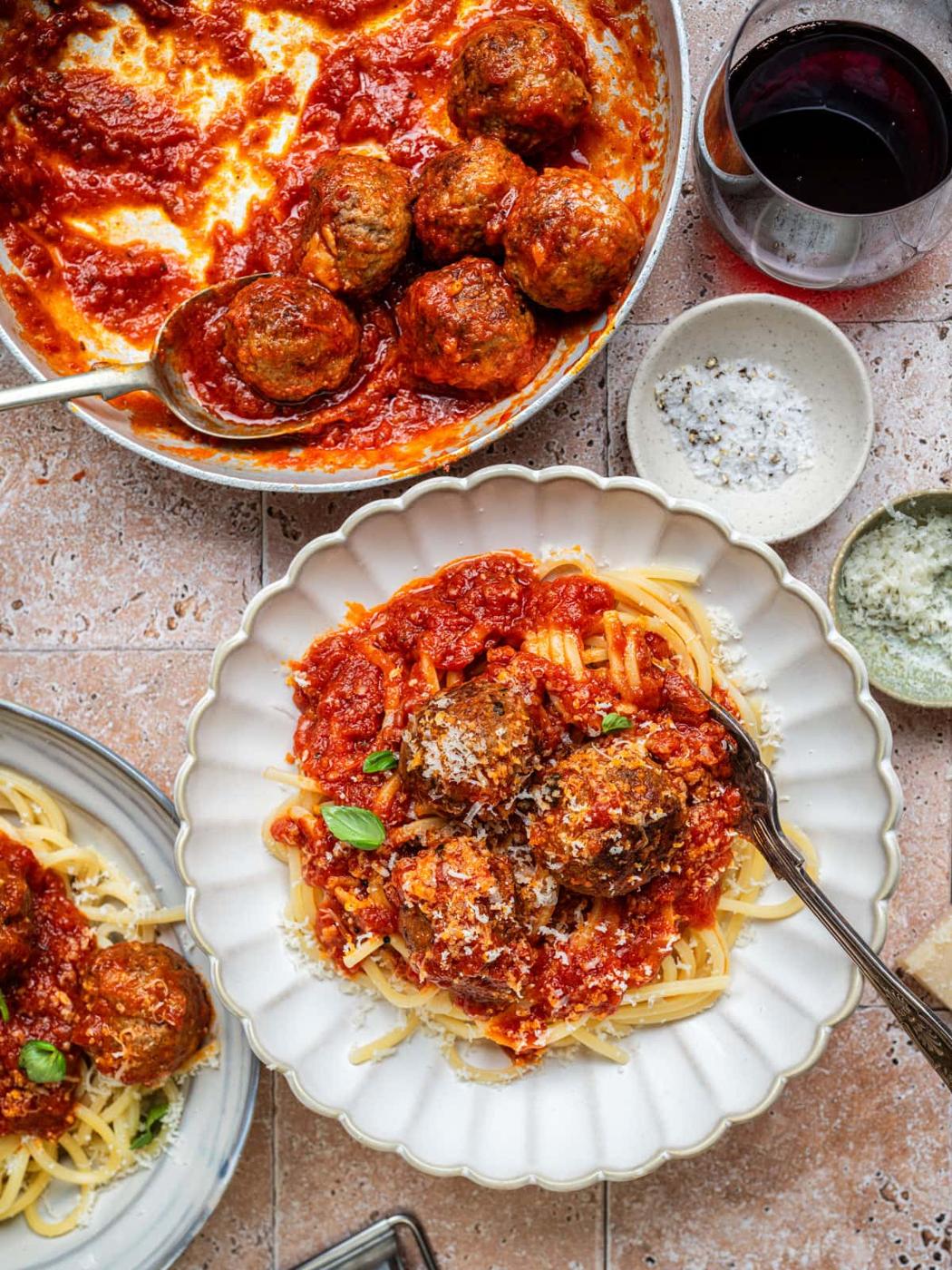 A plate of spaghetti topped with meatballs in tomato sauce, garnished with basil, sits next to a pan of meatballs, a glass of red wine, and small bowls of grated cheese and salt on a tiled surface.
