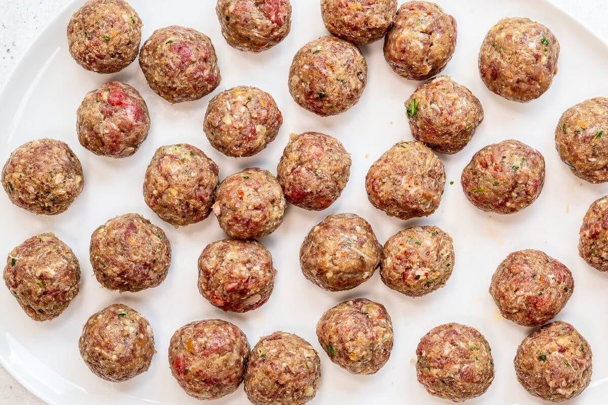 A white oval plate holds several evenly spaced, uncooked meatballs made with ground meat and herbs on a light-colored surface.