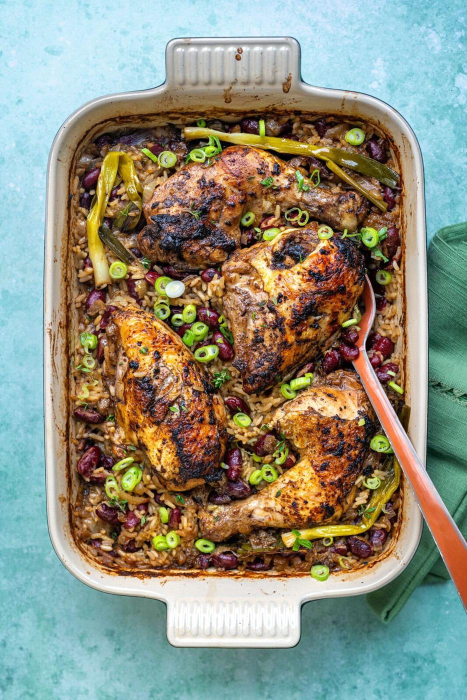 A baking dish filled with jerk chicken rice and peas features roasted seasoned chicken atop a bed of rice and kidney beans, garnished with sliced green onions and whole green chilies. A serving spoon rests in the dish.