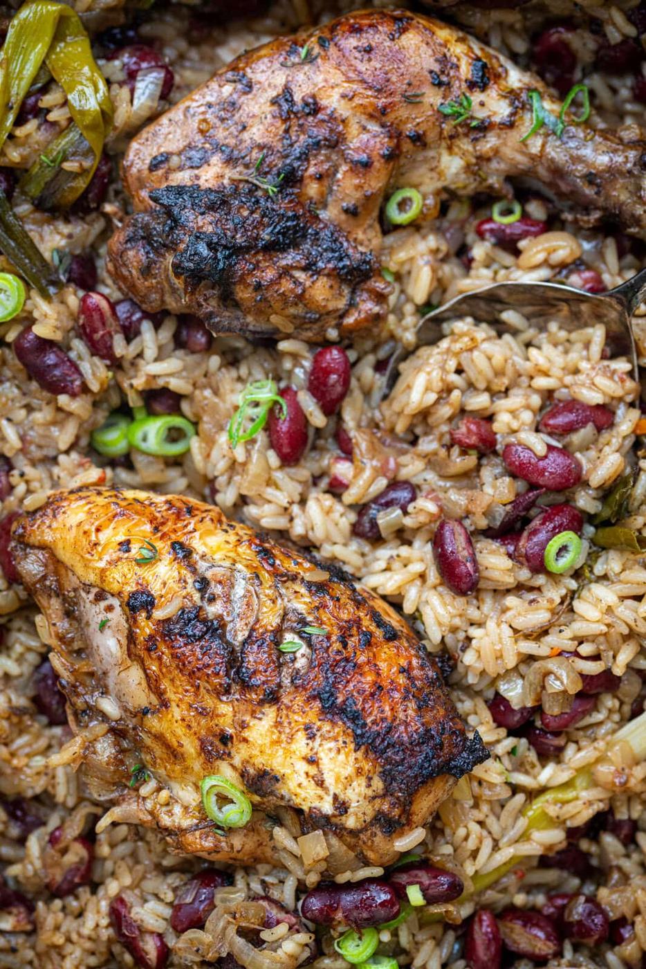 Two pieces of seasoned, roasted jerk chicken served on a bed of rice mixed with red kidney beans, garnished with chopped green onions. The dish looks flavorful and is cooked with herbs and spices.