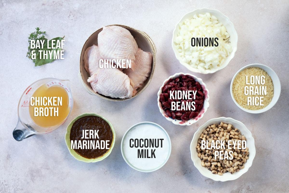 Top-down view of labeled ingredients in bowls and cups: bay leaf & thyme, chicken, onions, kidney beans, long grain rice, chicken broth, jerk marinade, coconut milk, and black eyed peas on a neutral surface.