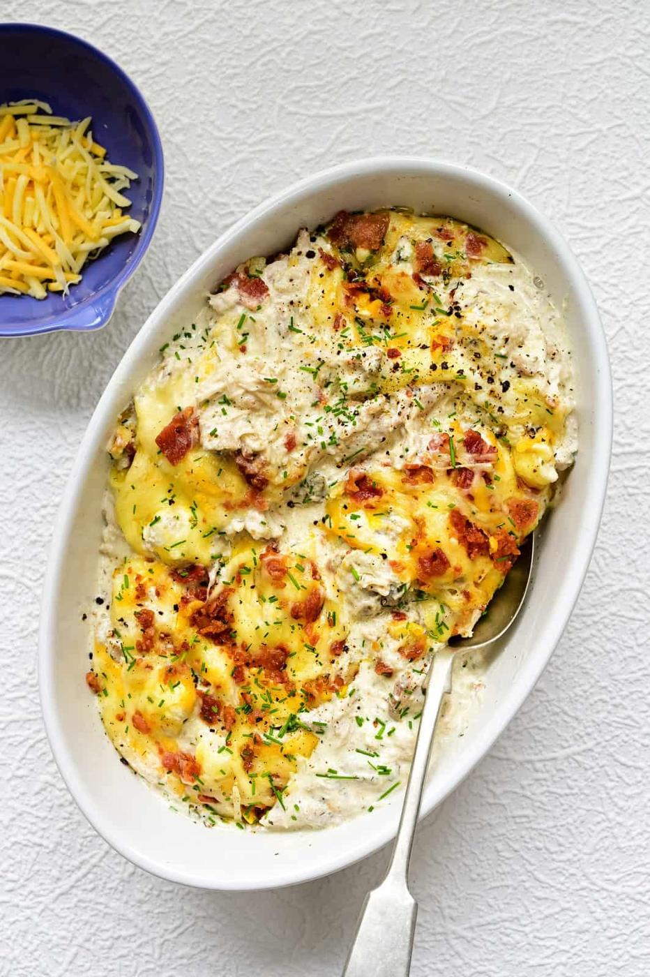 An oval white dish filled with creamy, cheesy Keto Crack Chicken casserole topped with melted cheese, herbs, and bacon bits, with a spoon inside. A small bowl of shredded cheese sits nearby on a textured white surface.
