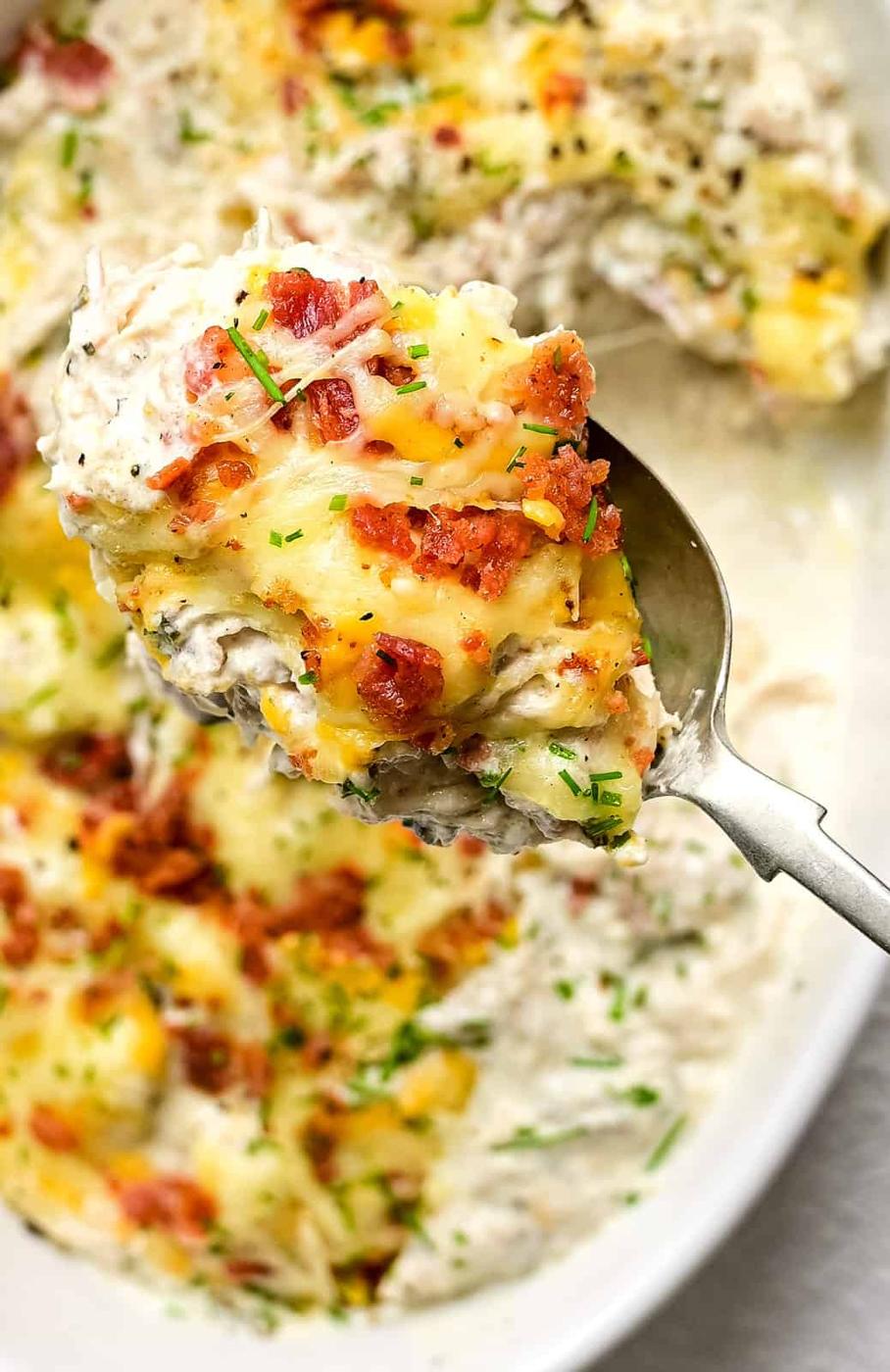 A close-up of a serving spoon holding a portion of creamy, keto crack chicken casserole topped with melted cheese, crispy bacon bits, and chopped herbs, with more casserole in a dish underneath.
