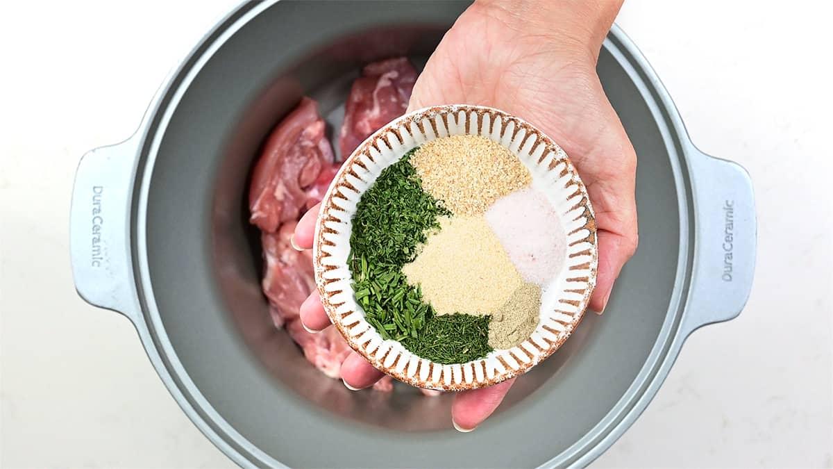 A hand holds a small bowl of mixed dried herbs and spices above a slow cooker filled with raw chicken pieces, ready to transform into flavorful keto crack chicken.