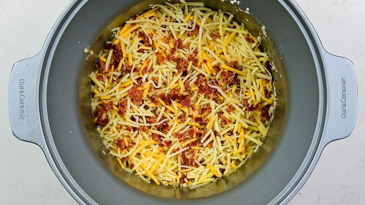 A slow cooker filled with keto crack chicken, featuring a savory mixture of shredded cheese and crumbled bacon, ready to be cooked.