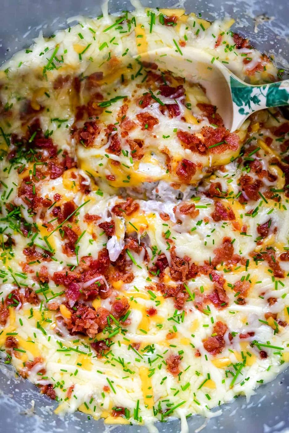 A close-up of creamy keto mashed potatoes topped with melted cheese, crispy bacon bits, and chopped chives, with a spoon scooping some from the dish—perfect comfort food for any slow cooker meal.