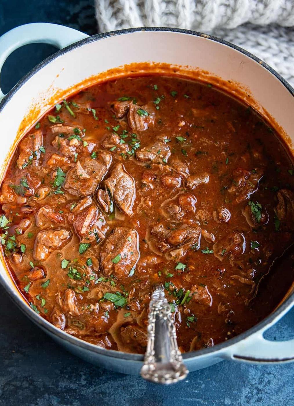 A pot filled with rich, chunky kokinisto beef stew in tomato sauce, garnished with chopped herbs, sits on a dark countertop with a spoon resting inside.