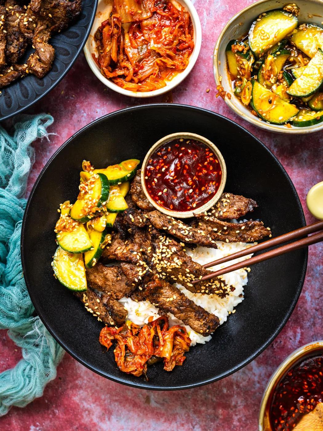 A Bulgogi bowl filled with white rice, Korean BBQ beef strips, sesame seeds, kimchi, cucumber slices, and a small bowl of red chili sauce. Surrounding dishes contain more kimchi and cucumbers, all on a pink surface.