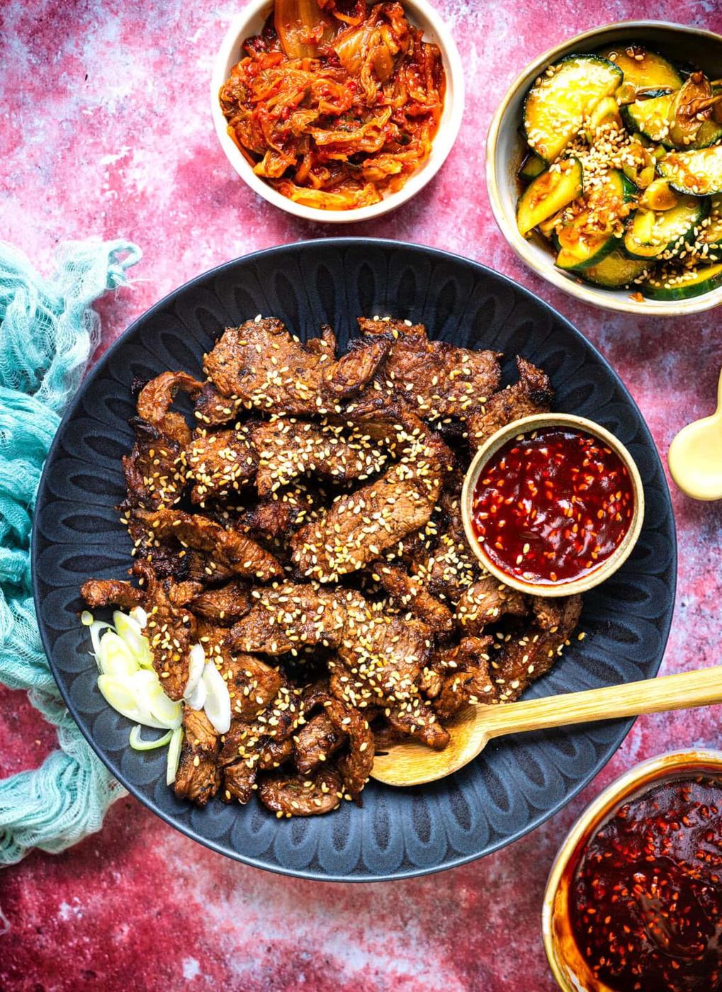 A black plate of sliced bulgogi beef topped with sesame seeds, garnished with green onions, with a small bowl of red dipping sauce, surrounded by dishes of kimchi and cucumber salad on a pink surface.