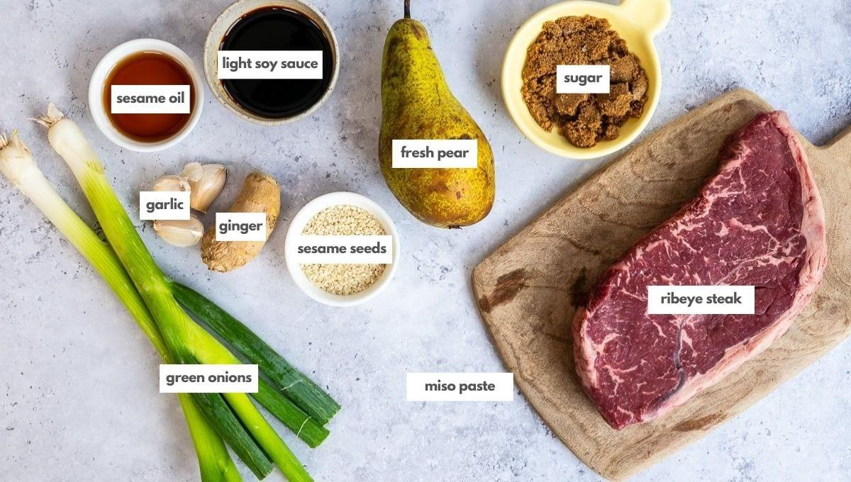 Top-down view of labeled ingredients on a light surface: green onions, garlic, ginger, sesame oil, light soy sauce, sesame seeds, fresh pear, miso paste, brown sugar, and a raw ribeye steak on a wooden board.