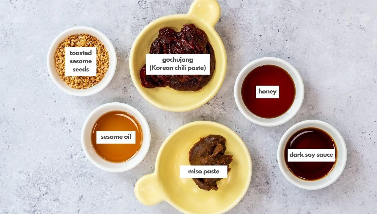 Five small bowls on a light surface containing labeled ingredients: toasted sesame seeds, gochujang (Korean chili paste), honey, sesame oil, miso paste, and dark soy sauce.